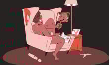 A brown-skinned woman sits naked in an arm chair holding a mirror in front of herself as she pleasures herself.