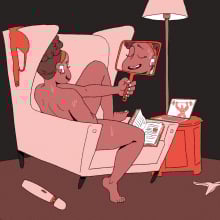 A brown-skinned woman sits naked in an arm chair holding a mirror in front of herself as she pleasures herself.