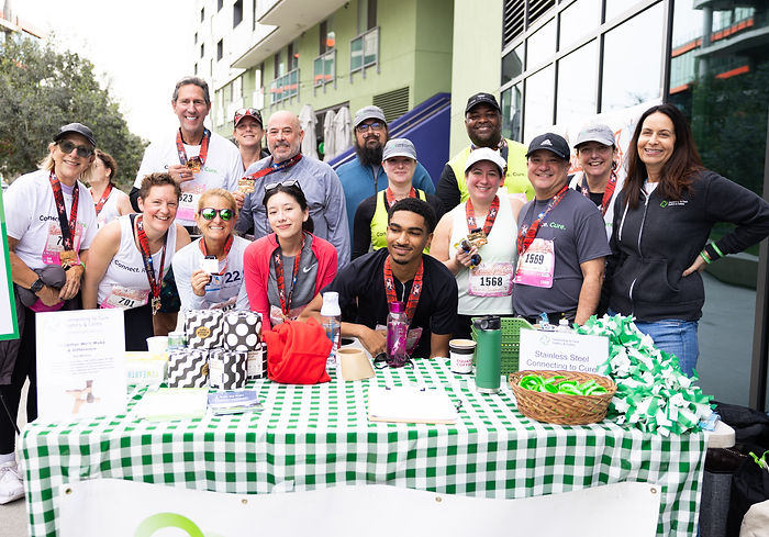 A group of people, many wearing race bibs and medals, gather smiling behind an information table covered in a green checkered cloth at an outdoor event. The table has a basket of green wristbands, water bottles, gifts, and informational signs. The group appears to be celebrating post-race and supporting an organization called Connecting to Cure Crohn’s and Colitis.